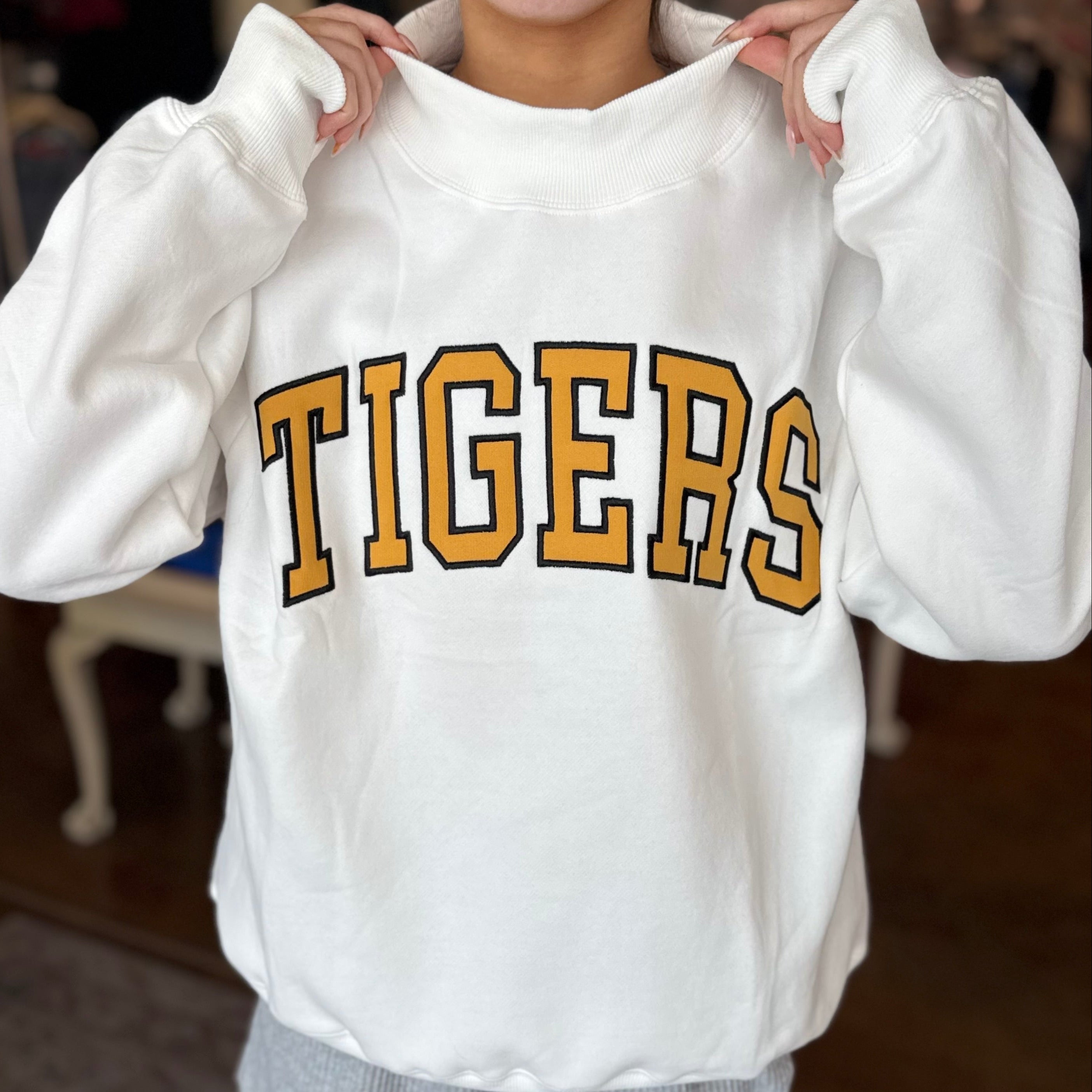 A person wearing a white sweatshirt with 'TIGERS' printed across the chest in gold and black varsity-style lettering, holding the collar open. The background shows a blurred indoor room with wooden floors and furniture.