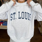 A person wearing an oversized light heather gray sweatshirt with 'ST. LOUIS' printed across the chest in dark navy collegiate lettering, paired with gray shorts in a store setting.