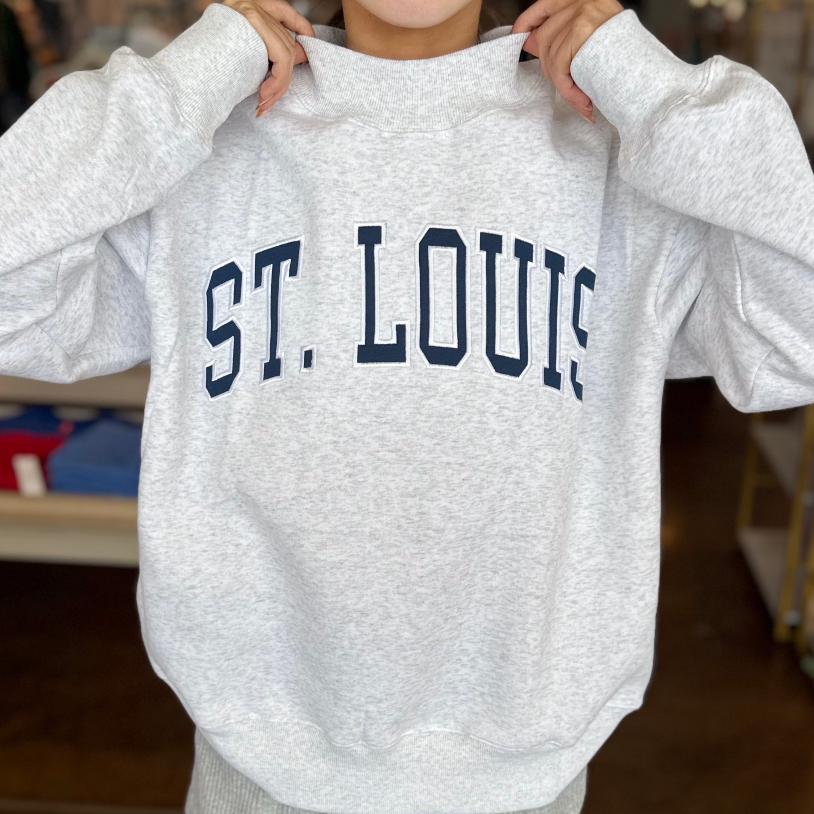 A person wearing an oversized light heather gray sweatshirt with 'ST. LOUIS' printed across the chest in dark navy collegiate lettering, paired with gray shorts in a store setting.