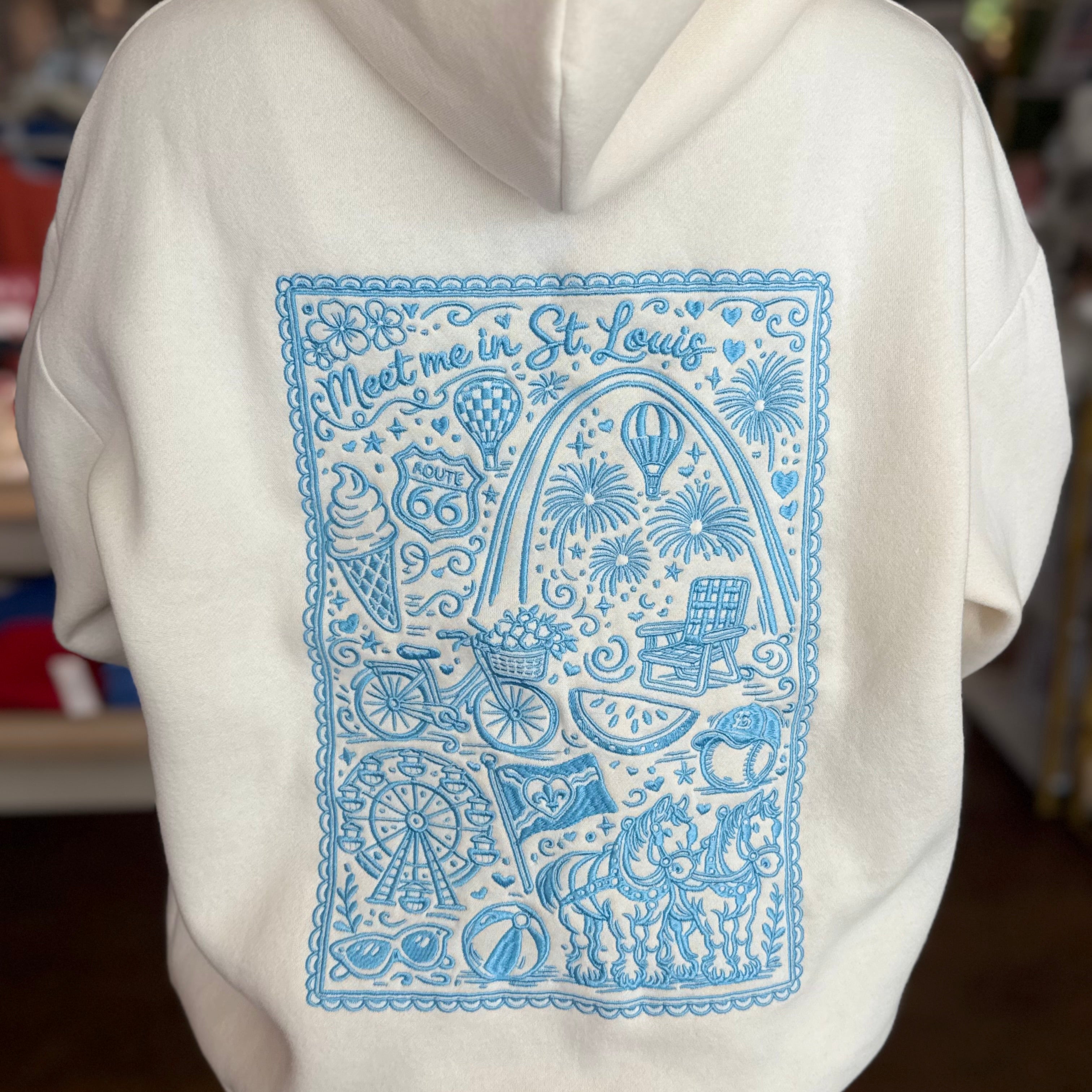 A person wearing a white hoodie with a large light blue graphic print on the back featuring illustrations and the text 'Meet me in St. Louis'.
