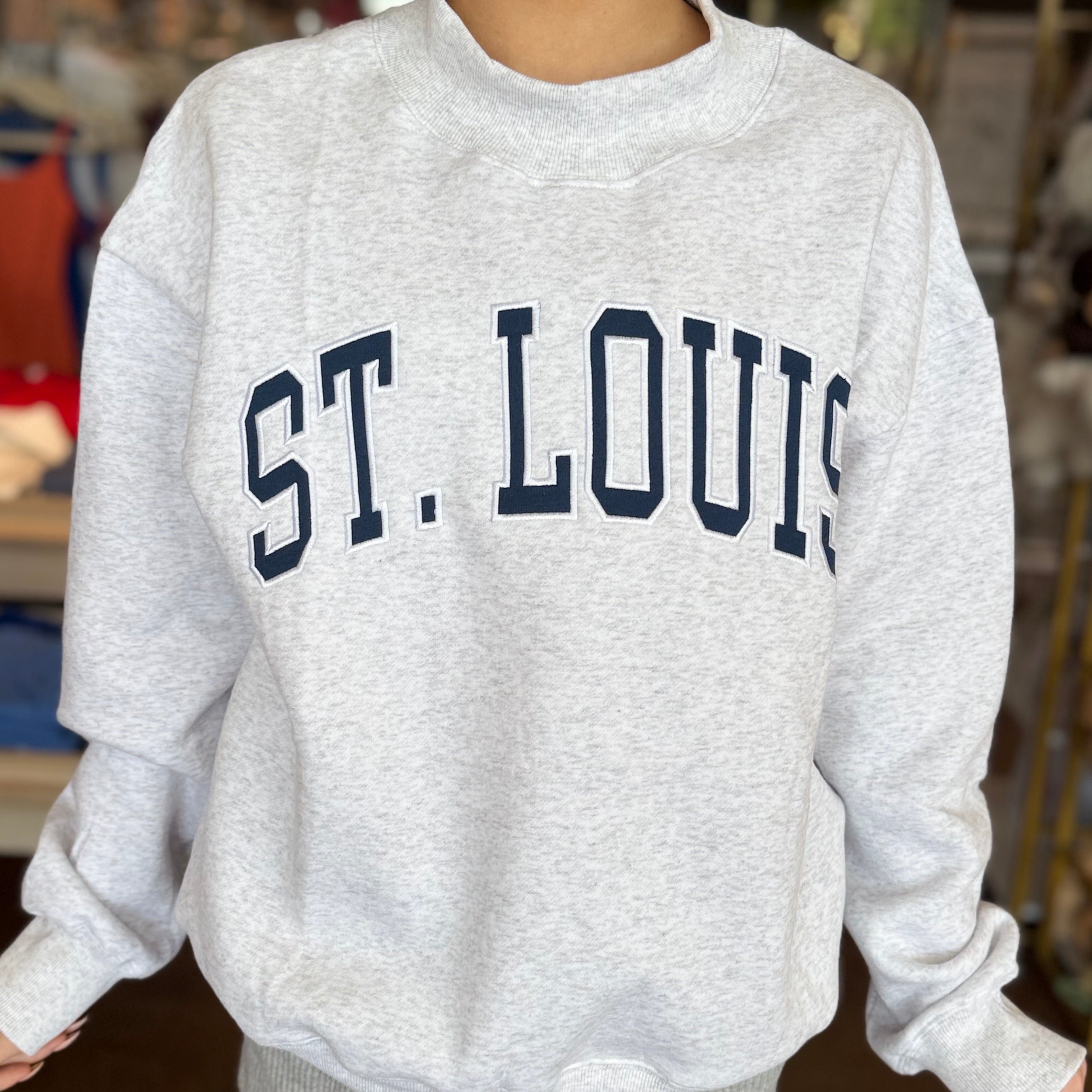 A person wearing a light heather gray crewneck sweatshirt with 'ST. LOUIS' printed across the chest in dark navy collegiate lettering, photographed in a store setting.